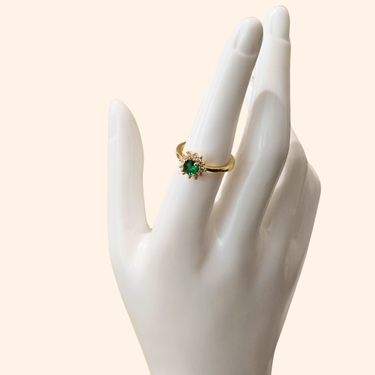 The Emerald Flower Gold Ring by Roots Jewellery features a stunning green emerald at its center, accented by dazzling clear stones. Its sophisticated design is set on a simple backdrop and includes an adjustable feature for the ideal fit.