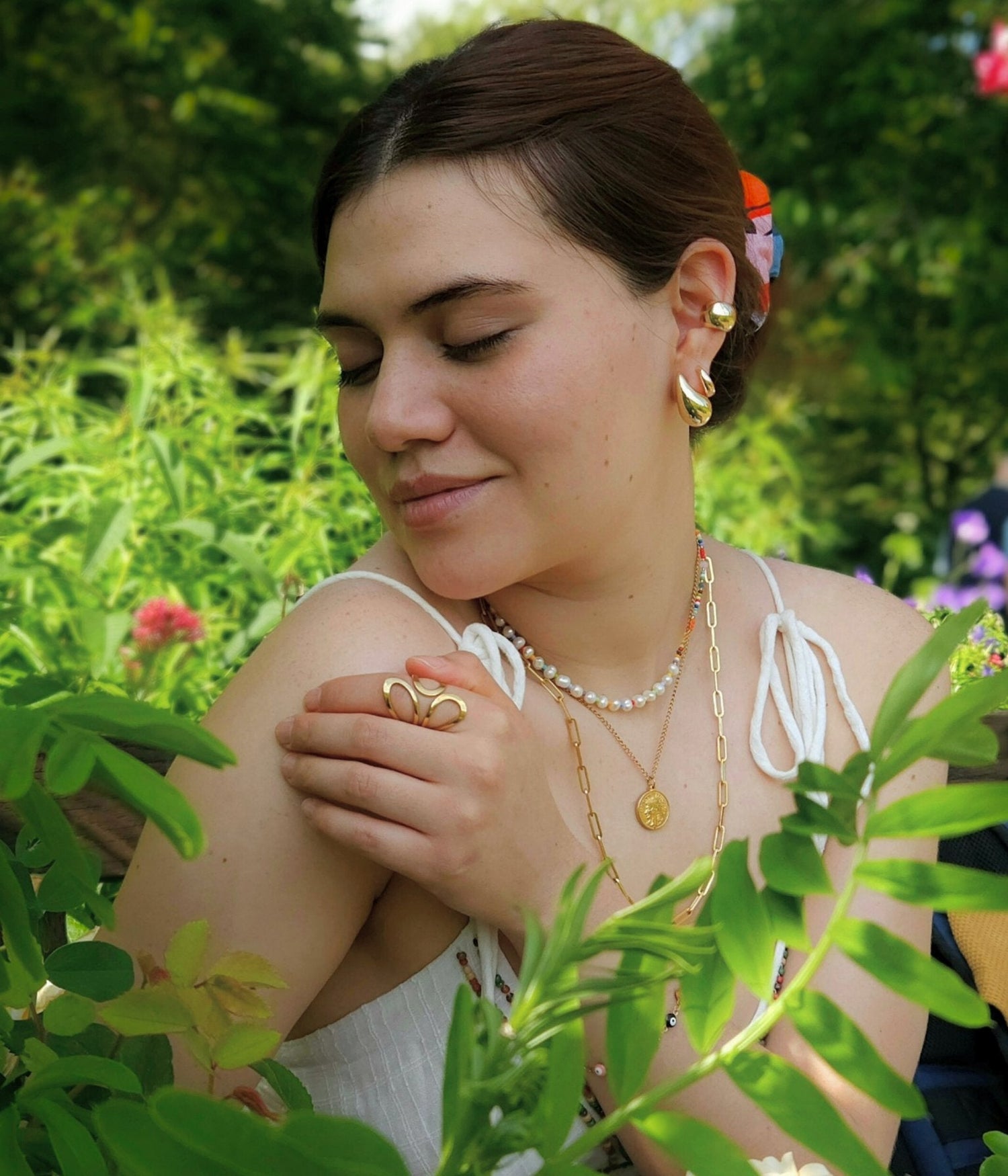 A woman in a garden wearing gold jewelry and a white dress, eyes closed, gently touching her shoulder.