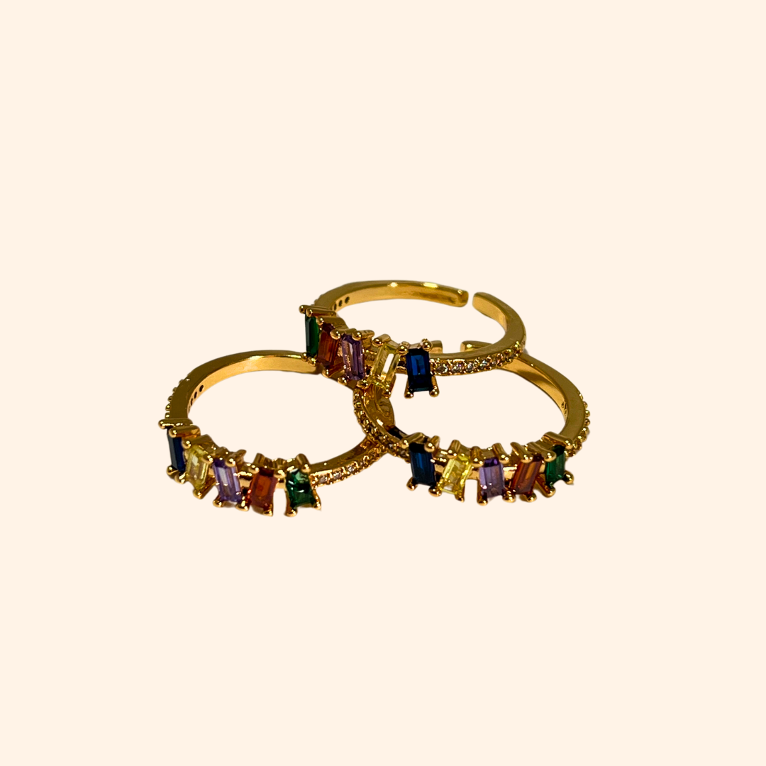 The Five Gems Gold Ring by Roots Jewellery features a dazzling arrangement of five rectangular zircons in vibrant hues—red, green, purple, blue, and clear—set diagonally.