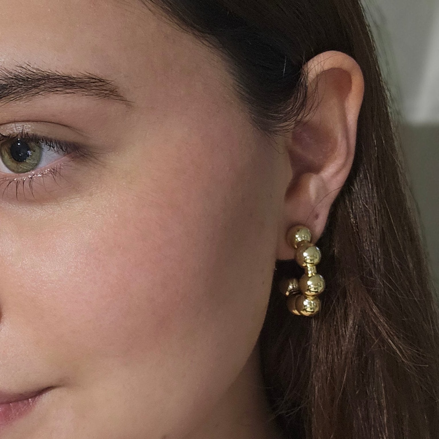 Close-up of a person's ear adorned with stylish Roots Jewellery hoops, showcasing Gold Ball Hoops Earrings. A portion of the face is visible, highlighting the jewelry's elegance with its gold and rhodium plating.