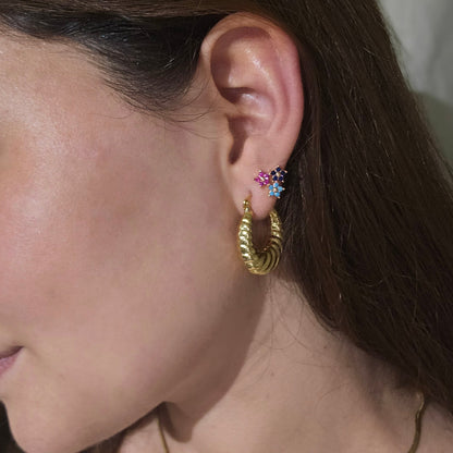 A woman with long brown hair is wearing a simple gold necklace and colorful Triple Flower Gold Earrings by Roots Jewellery, looking to the side.