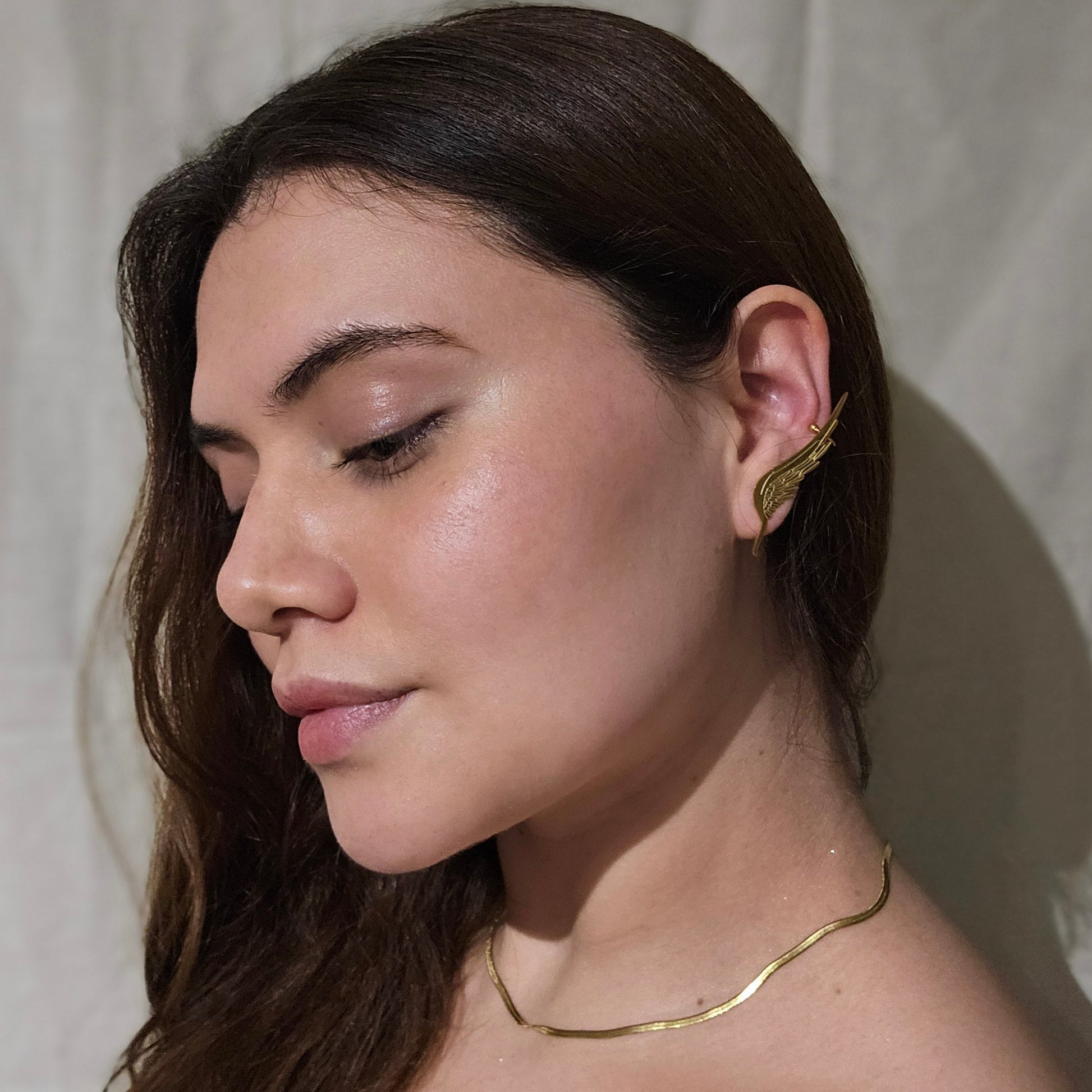 White ceramic ear model adorned with an elegant gold wing-shaped ear cuff, inspired by the Golden Angel Wings Earrings from Roots Jewellery, displayed on a beige background.