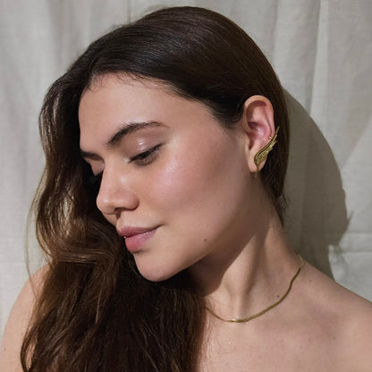 White ceramic ear model adorned with an elegant gold wing-shaped ear cuff, inspired by the Golden Angel Wings Earrings from Roots Jewellery, displayed on a beige background.