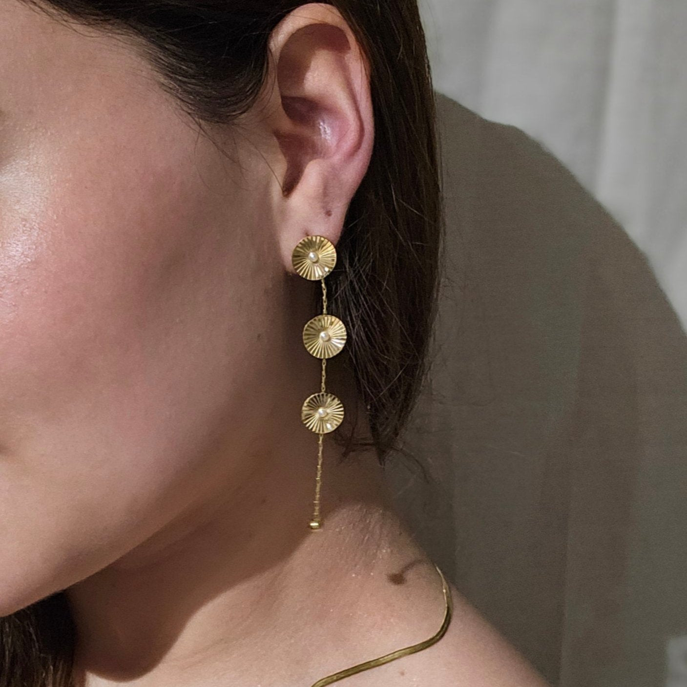 The Cosmic Earrings by Roots Jewellery feature silver and gold drop designs with circular patterns and elongated elements, providing a contemporary appeal inspired by cosmic motifs. They are showcased against a neutral backdrop.