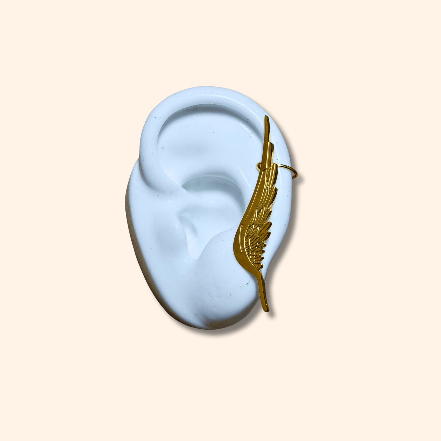 White ceramic ear model adorned with an elegant gold wing-shaped ear cuff, inspired by the Golden Angel Wings Earrings from Roots Jewellery, displayed on a beige background.