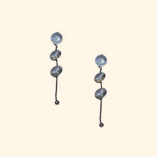 The Cosmic Earrings by Roots Jewellery feature silver and gold drop designs with circular patterns and elongated elements, providing a contemporary appeal inspired by cosmic motifs. They are showcased against a neutral backdrop.