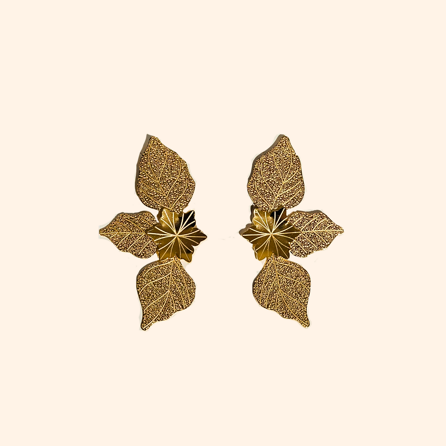 The Golden Leaf Charm Earrings by Roots Jewellery, showcasing intricate leaf designs, rest gracefully on a light beige surface and are equipped with durable stainless steel backings for enduring sophistication.