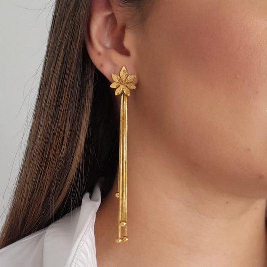 A woman wearing Roots Jewellery's Leaf Cascade Earrings showcases a long gold piece adorned with an intricate floral design at the top.