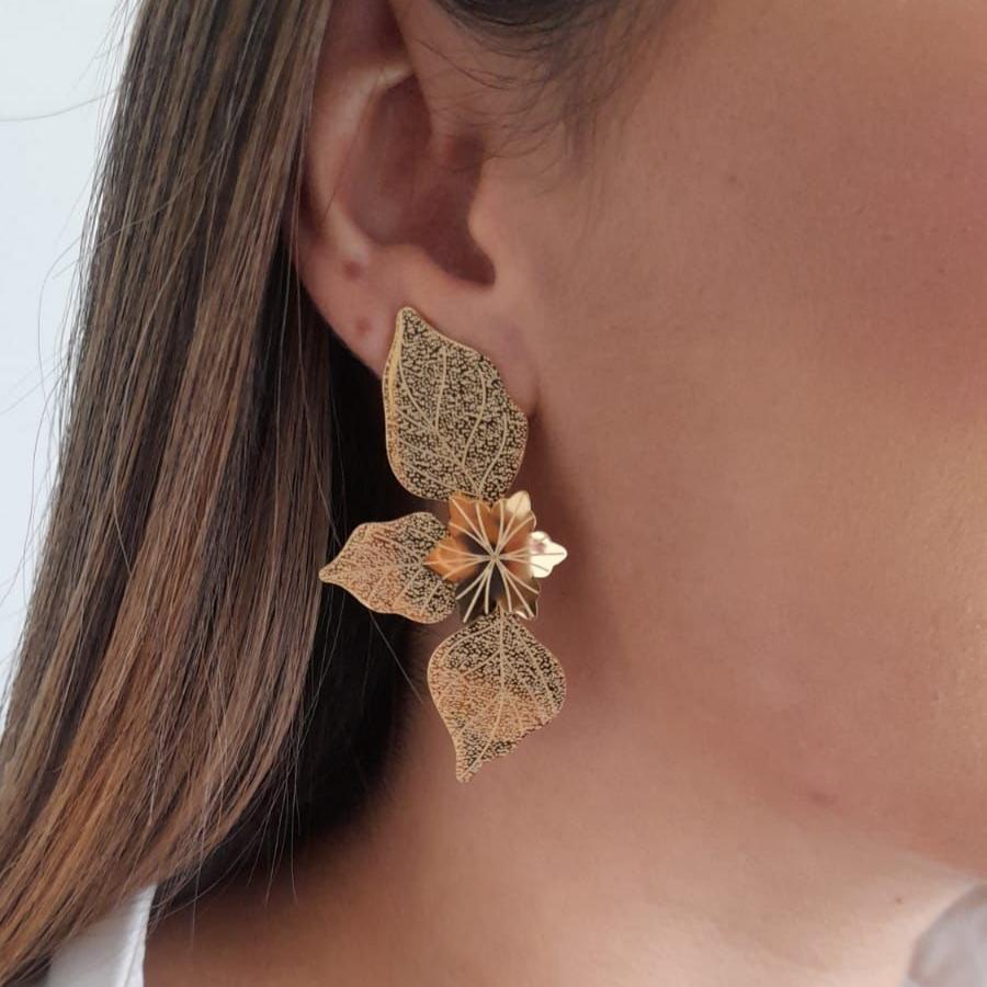 The Golden Leaf Charm Earrings by Roots Jewellery, showcasing intricate leaf designs, rest gracefully on a light beige surface and are equipped with durable stainless steel backings for enduring sophistication.