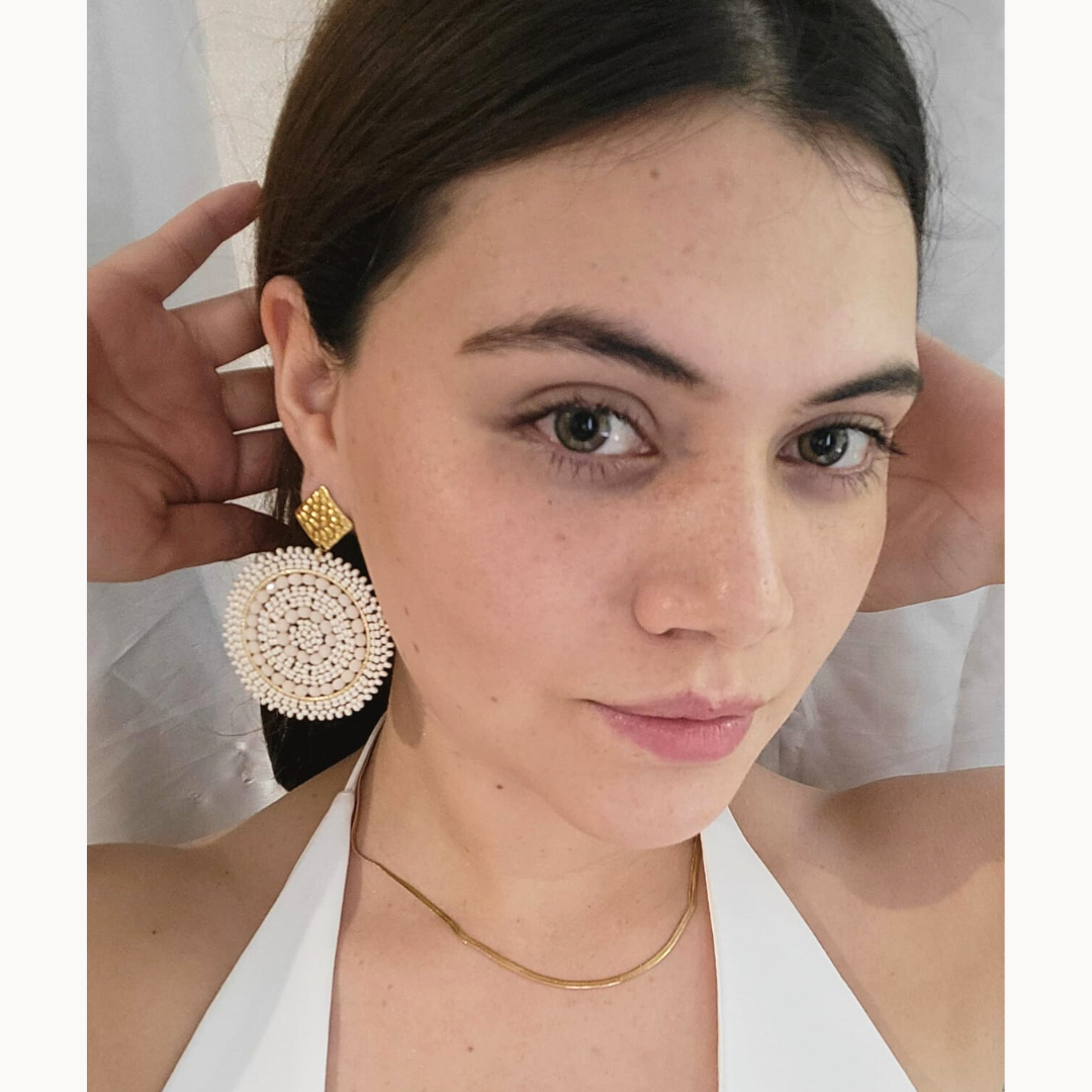 A woman poses elegantly wearing the Radiant Spiral Beaded Earrings by Roots Jewellery, featuring intricate beadwork, along with a matching gold necklace.